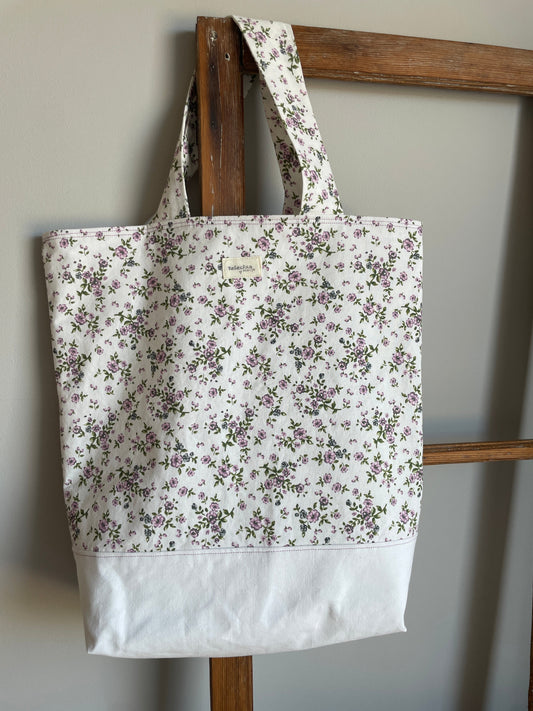 Tygväska med vit botten och vit ovandel med små lila blommor, två handtag och hängande på en gammal fönsterram.