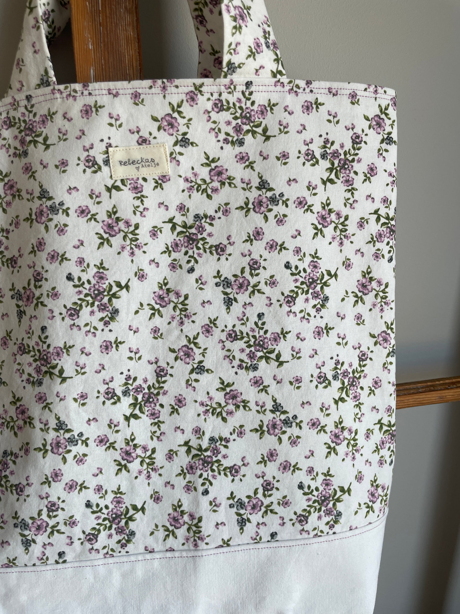 Tygväska med vit botten och vit ovandel med små lila blommor, två handtag och hängande på en gammal fönsterram.