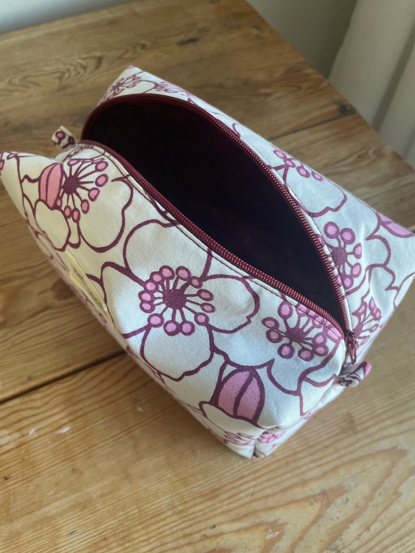 Blommig boxnecessär med stora vita och rosa blommor, vinröd dragkedja och vinrödrosa innerfoder.