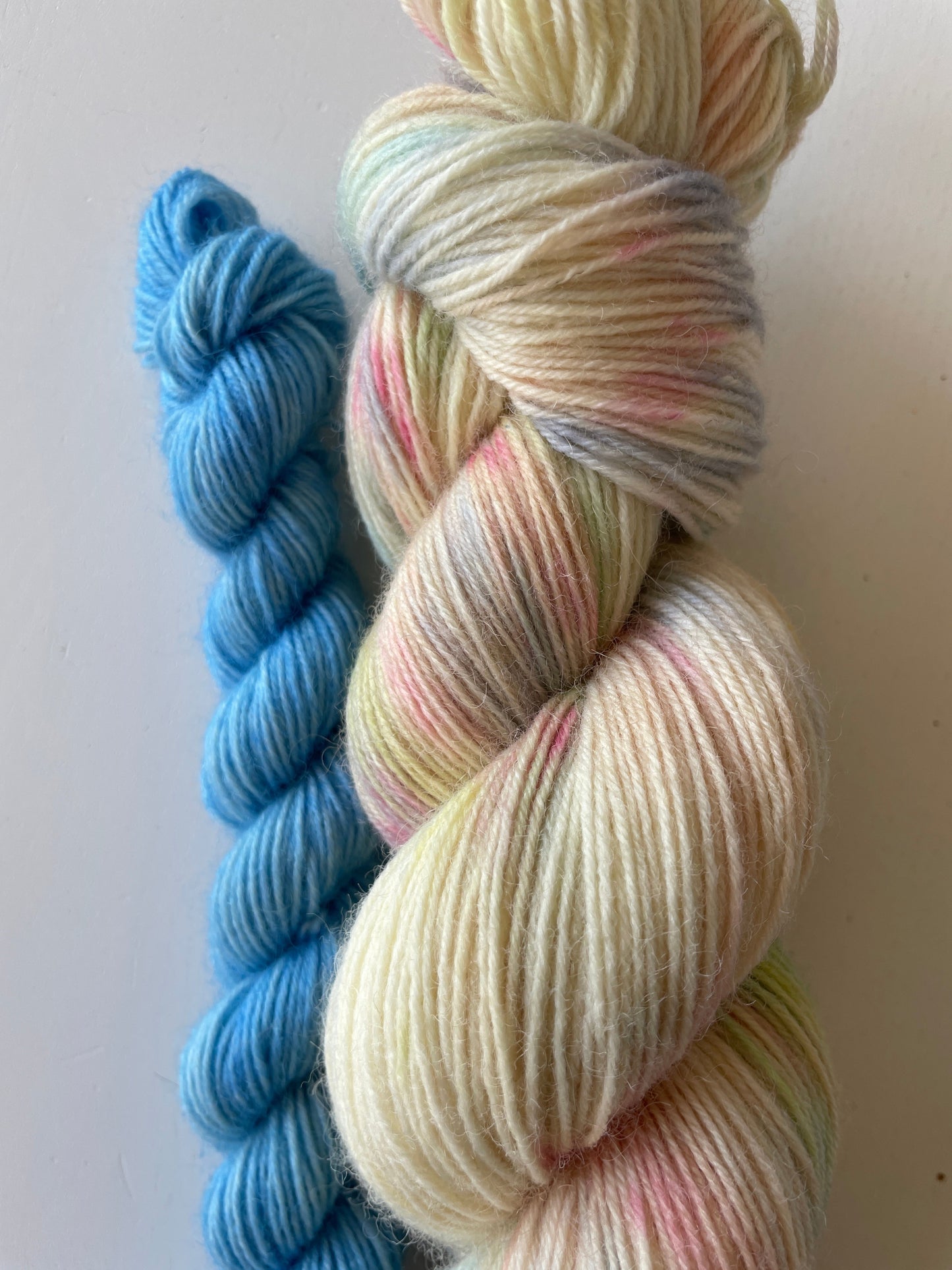 Handfärgat melerat garnnystan på 100 g i vitt och beige med färgstänk i gul, blå, rosa och grön samt ett blått miniskein på 20 g.