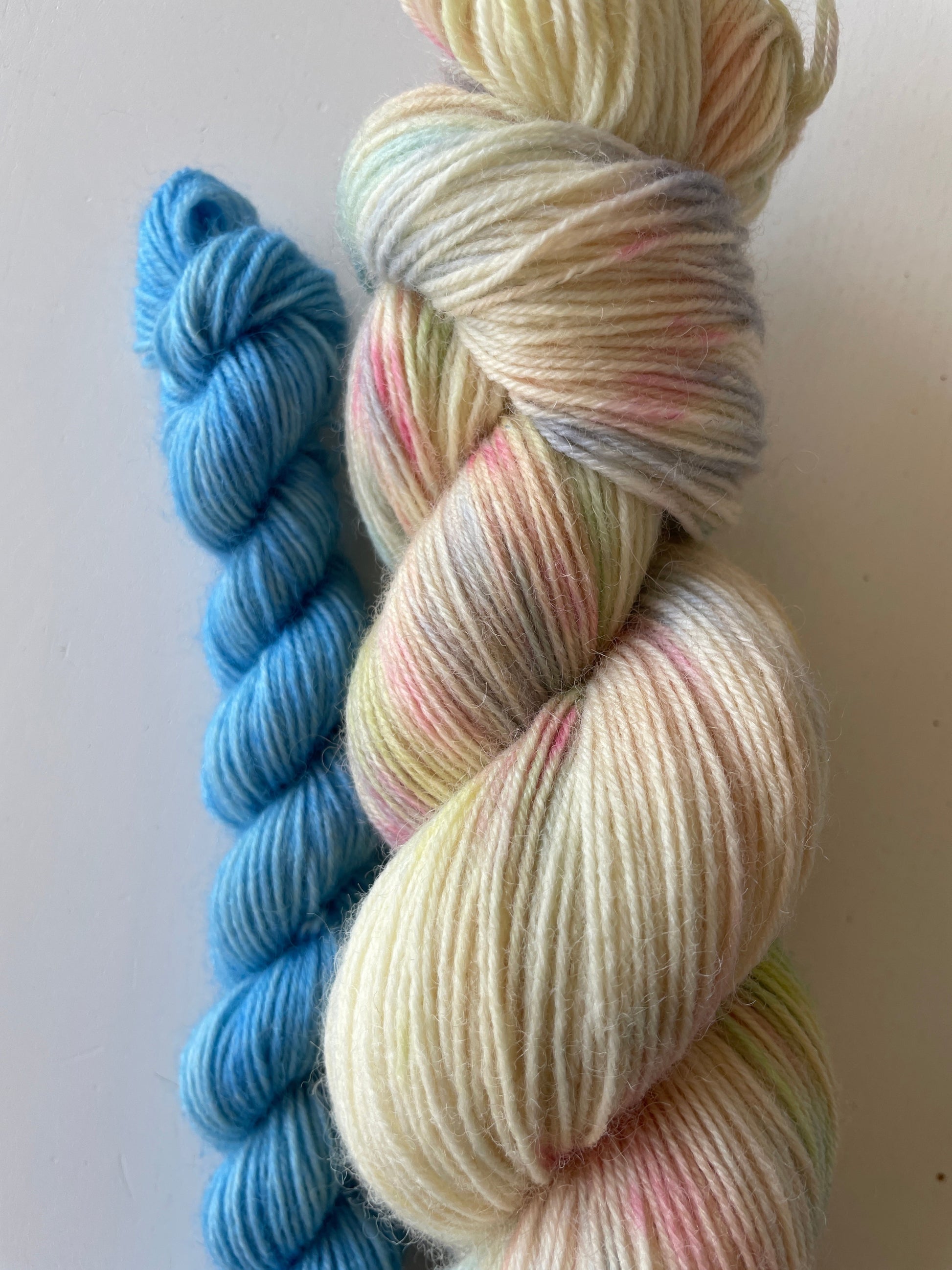 Handfärgat melerat garnnystan på 100 g i vitt och beige med färgstänk i gul, blå, rosa och grön samt ett blått miniskein på 20 g.