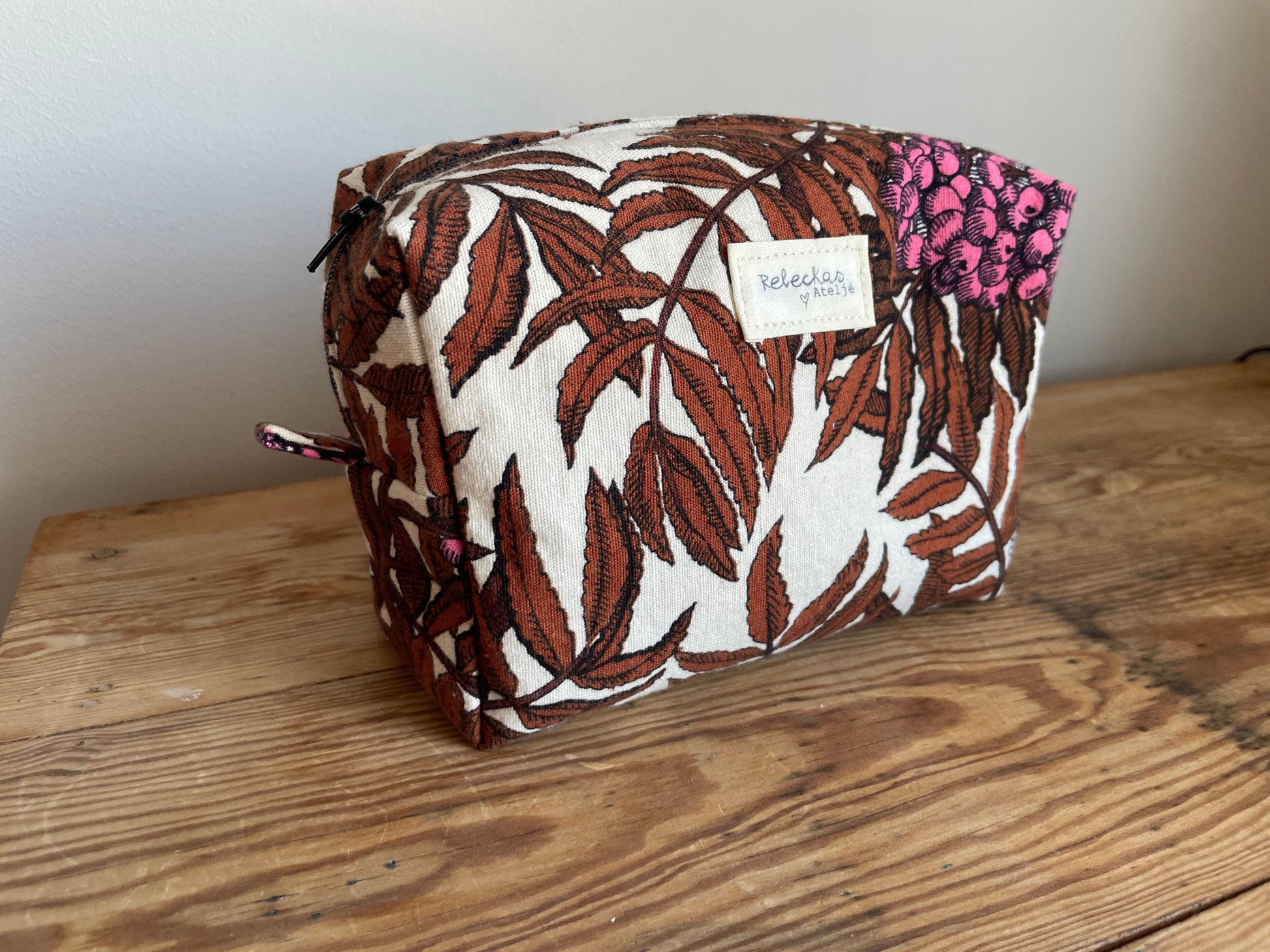 Handgjord boxnecessär i tyg med beige bakgrund, mörkbruna löv, starkt rosa bärklasar och dragkedja.