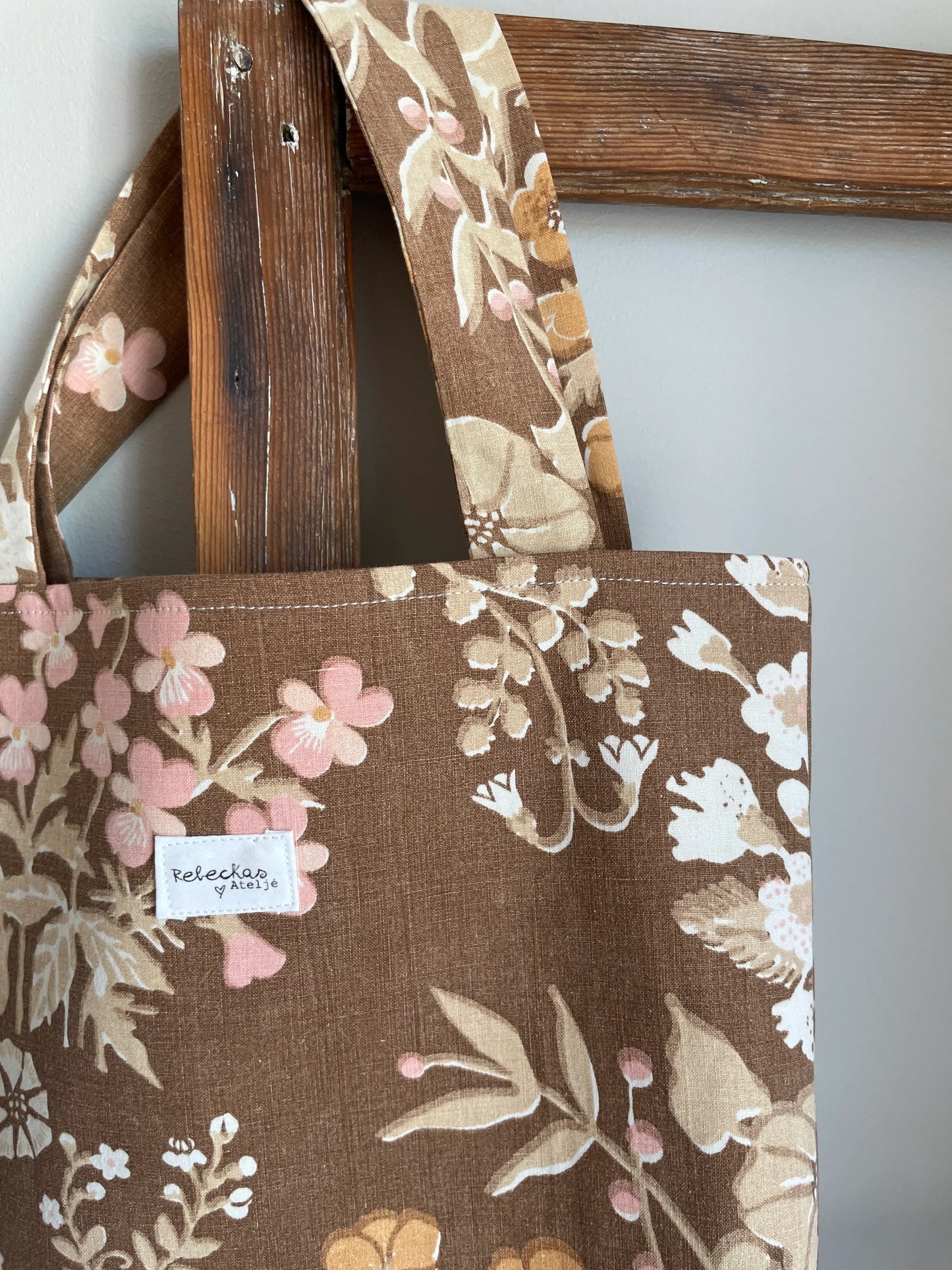 Handsydd projektväska i brun bomull med vintageinspirerat blommönster i rosa, beige, creme och honungsgula toner. Praktisk tygväska med långa handtag för stickning, virkning och garnförvaring. Väskan hänger på en rustik trästege mot en ljus vägg och passar perfekt som projektpåse för handarbete och kreativa projekt.
