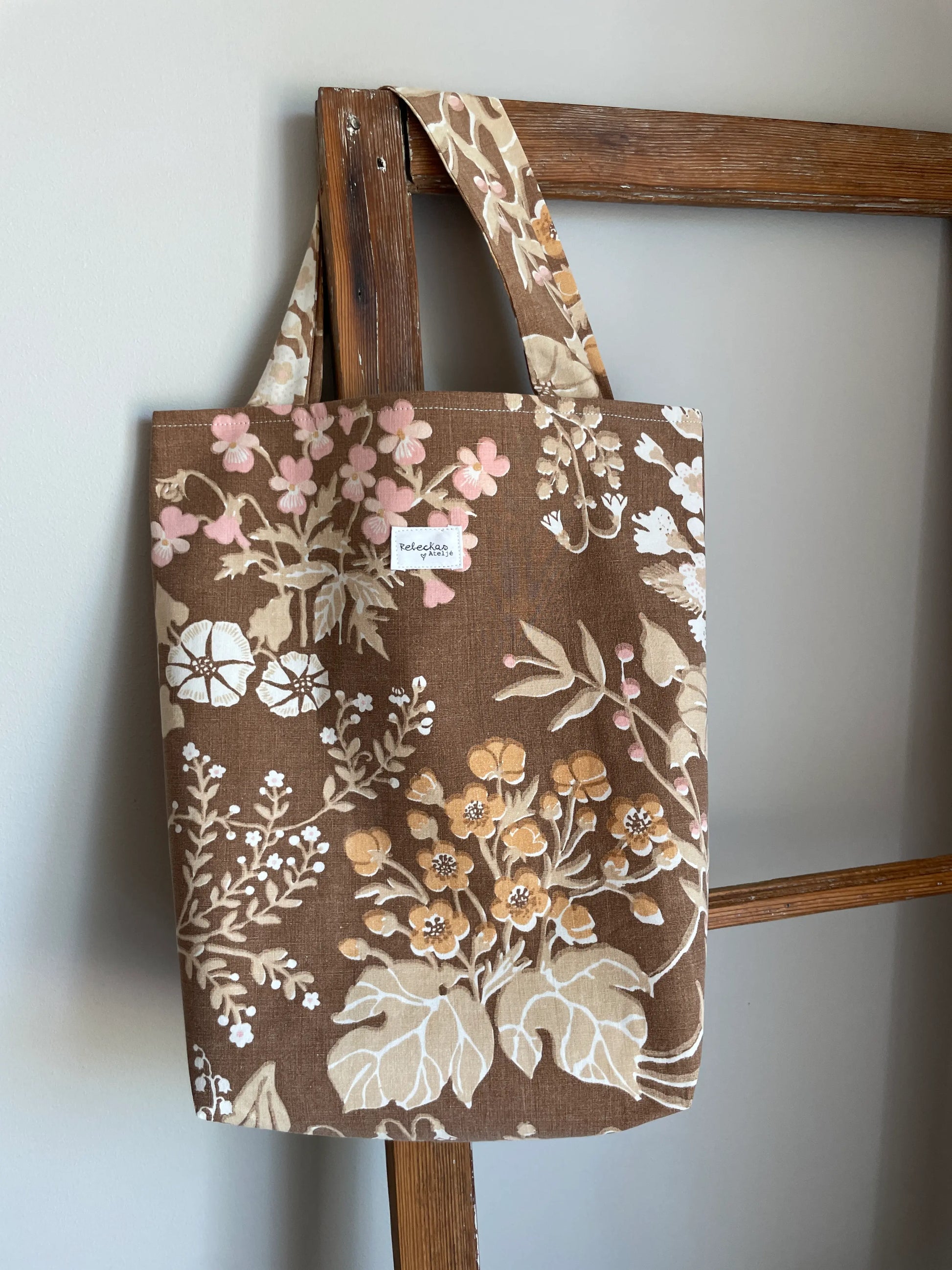Handsydd projektväska i brun bomull med vintageinspirerat blommönster i rosa, beige, creme och honungsgula toner. Praktisk tygväska med långa handtag för stickning, virkning och garnförvaring. Väskan hänger på en rustik trästege mot en ljus vägg och passar perfekt som projektpåse för handarbete och kreativa projekt.