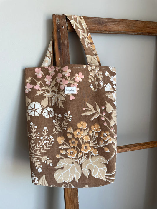 Handsydd projektväska i brun bomull med vintageinspirerat blommönster i rosa, beige, creme och honungsgula toner. Praktisk tygväska med långa handtag för stickning, virkning och garnförvaring. Väskan hänger på en rustik trästege mot en ljus vägg och passar perfekt som projektpåse för handarbete och kreativa projekt.