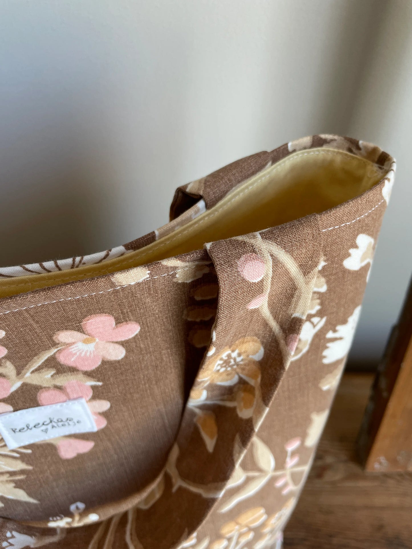 Handsydd projektväska i brun bomull med vintageinspirerat blommönster i rosa, beige, creme och honungsgula toner. Praktisk tygväska med långa handtag för stickning, virkning och garnförvaring. Väskan hänger på en rustik trästege mot en ljus vägg och passar perfekt som projektpåse för handarbete och kreativa projekt.