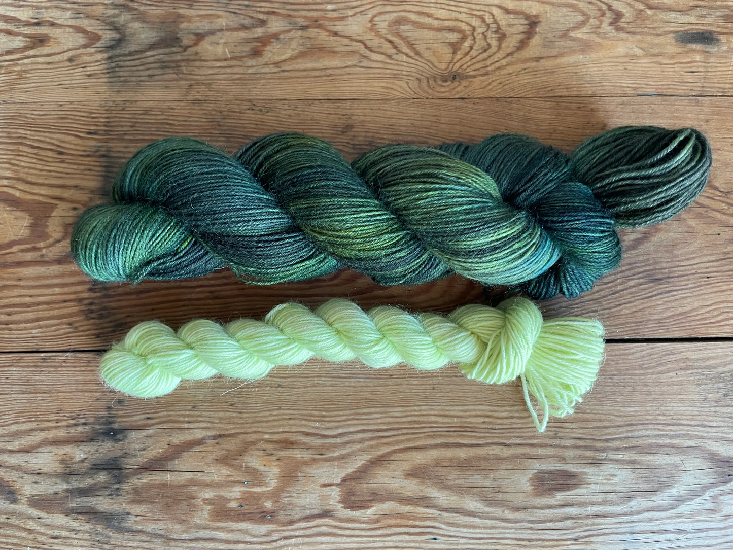 Handfärgat sockgarn-set med 100 g sockgarn i skogsgröna, petrolblå och mossgröna nyanser samt ett 20 g mini-nystan i ljus limegrön kontrastfärg. Garnset för stickade strumpor, sockor och accessoarer där mini-nystanet passar till häl, tå och mudd. Fotograferat på rustikt träbord som framhäver garnets färgskiftningar och glans.