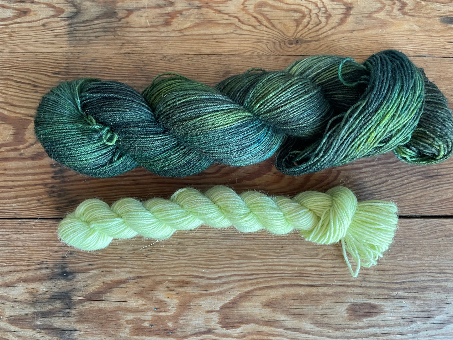 Handfärgat sockgarn-set med 100 g sockgarn i skogsgröna, petrolblå och mossgröna nyanser samt ett 20 g mini-nystan i ljus limegrön kontrastfärg. Garnset för stickade strumpor, sockor och accessoarer där mini-nystanet passar till häl, tå och mudd. Fotograferat på rustikt träbord som framhäver garnets färgskiftningar och glans.