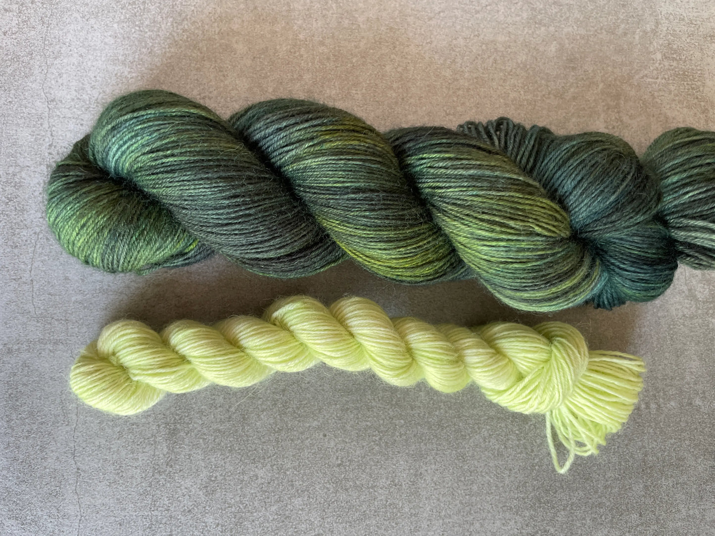 Handfärgat sockgarn-set med 100 g sockgarn i skogsgröna, petrolblå och mossgröna nyanser samt ett 20 g mini-nystan i ljus limegrön kontrastfärg. Garnset för stickade strumpor, sockor och accessoarer där mini-nystanet passar till häl, tå och mudd. Fotograferat på rustikt träbord som framhäver garnets färgskiftningar och glans.