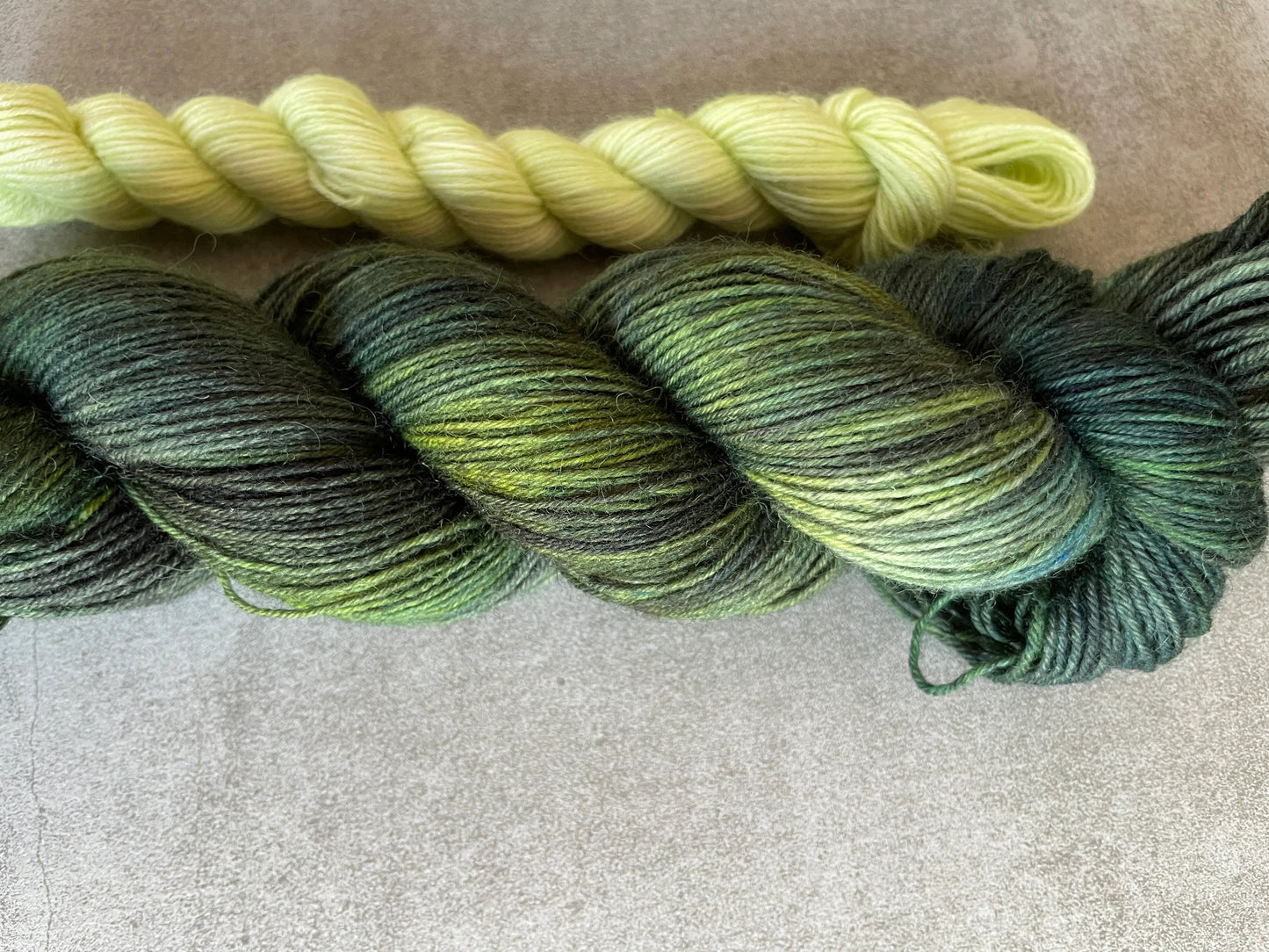 Handfärgat sockgarn-set med 100 g sockgarn i skogsgröna, petrolblå och mossgröna nyanser samt ett 20 g mini-nystan i ljus limegrön kontrastfärg. Garnset för stickade strumpor, sockor och accessoarer där mini-nystanet passar till häl, tå och mudd. Fotograferat på rustikt träbord som framhäver garnets färgskiftningar och glans.