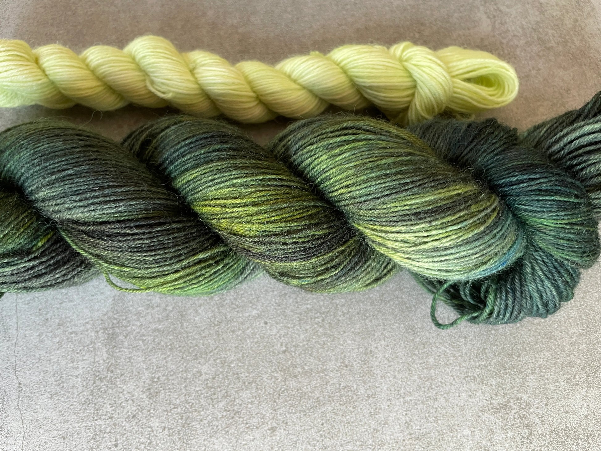 Handfärgat sockgarn-set med 100 g sockgarn i skogsgröna, petrolblå och mossgröna nyanser samt ett 20 g mini-nystan i ljus limegrön kontrastfärg. Garnset för stickade strumpor, sockor och accessoarer där mini-nystanet passar till häl, tå och mudd. Fotograferat på rustikt träbord som framhäver garnets färgskiftningar och glans.