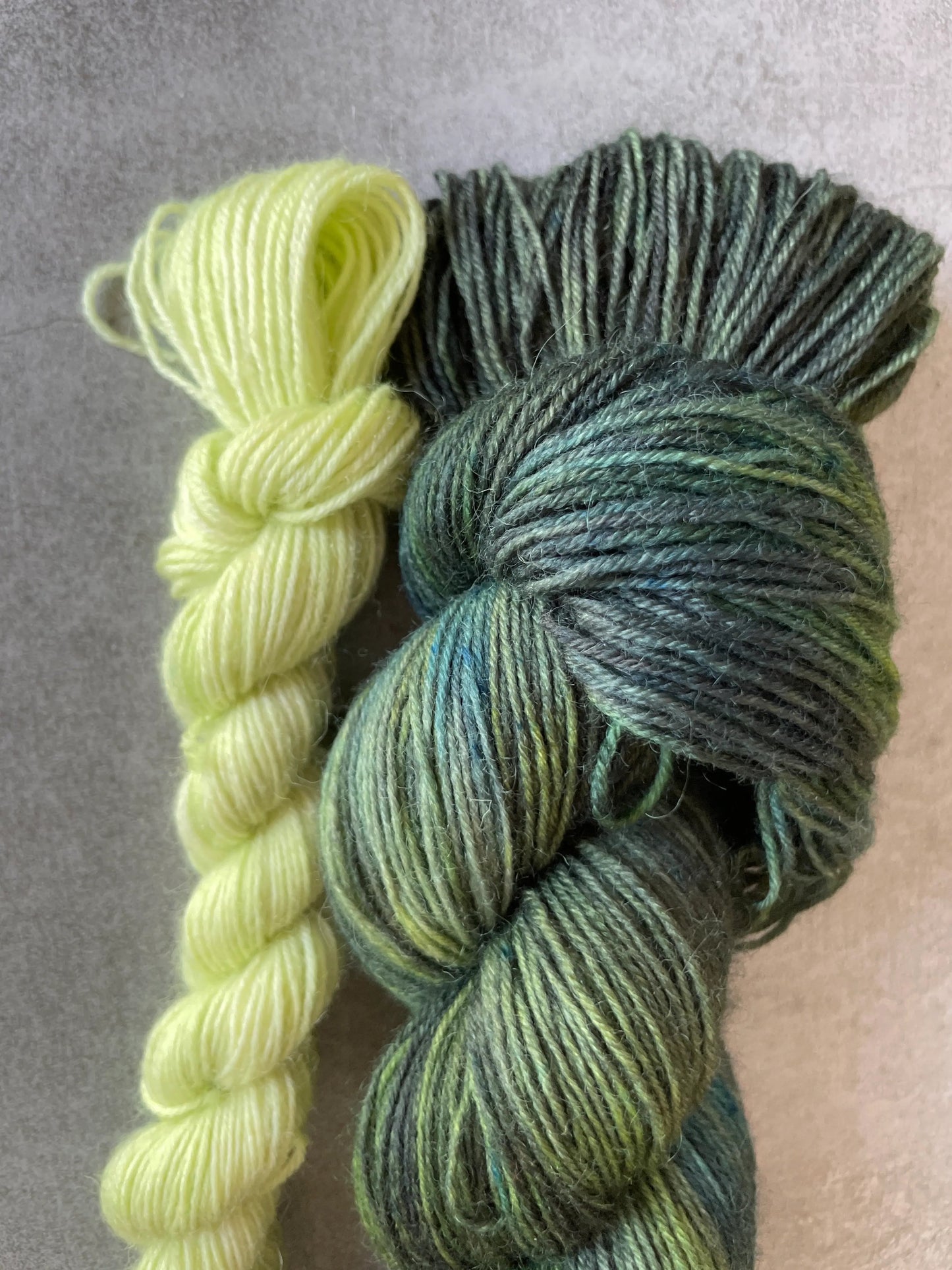 Handfärgat sockgarn-set med 100 g sockgarn i skogsgröna, petrolblå och mossgröna nyanser samt ett 20 g mini-nystan i ljus limegrön kontrastfärg. Garnset för stickade strumpor, sockor och accessoarer där mini-nystanet passar till häl, tå och mudd. Fotograferat på rustikt träbord som framhäver garnets färgskiftningar och glans.