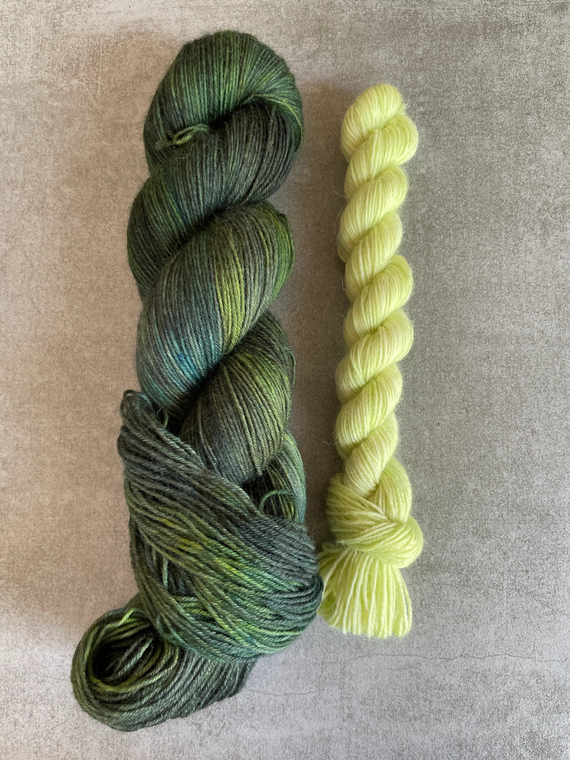 Handfärgat sockgarn-set med 100 g sockgarn i skogsgröna, petrolblå och mossgröna nyanser samt ett 20 g mini-nystan i ljus limegrön kontrastfärg. Garnset för stickade strumpor, sockor och accessoarer där mini-nystanet passar till häl, tå och mudd. Fotograferat på rustikt träbord som framhäver garnets färgskiftningar och glans.