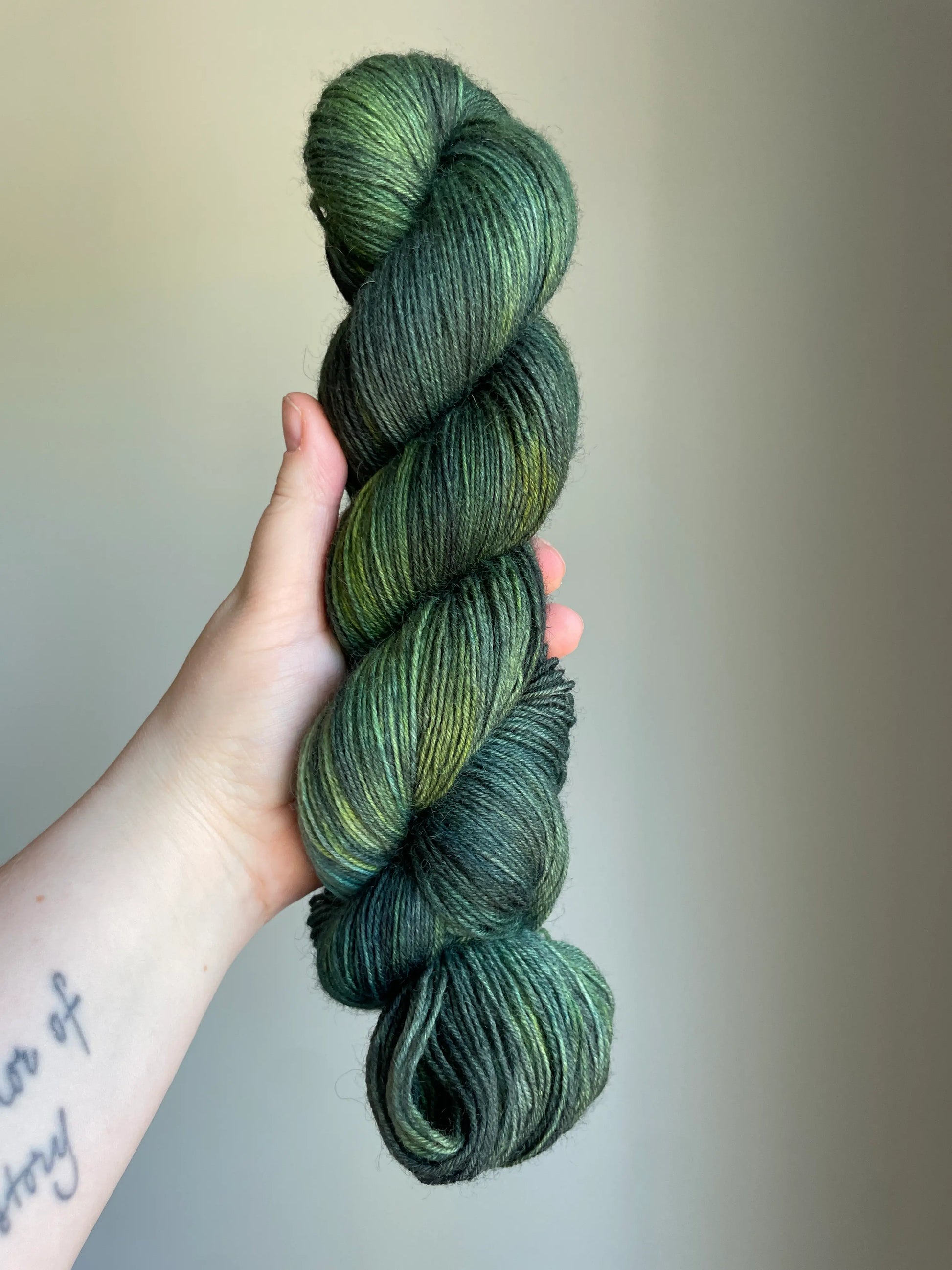 Handfärgat sockgarn-set med 100 g sockgarn i skogsgröna, petrolblå och mossgröna nyanser samt ett 20 g mini-nystan i ljus limegrön kontrastfärg. Garnset för stickade strumpor, sockor och accessoarer där mini-nystanet passar till häl, tå och mudd. Fotograferat på rustikt träbord som framhäver garnets färgskiftningar och glans.