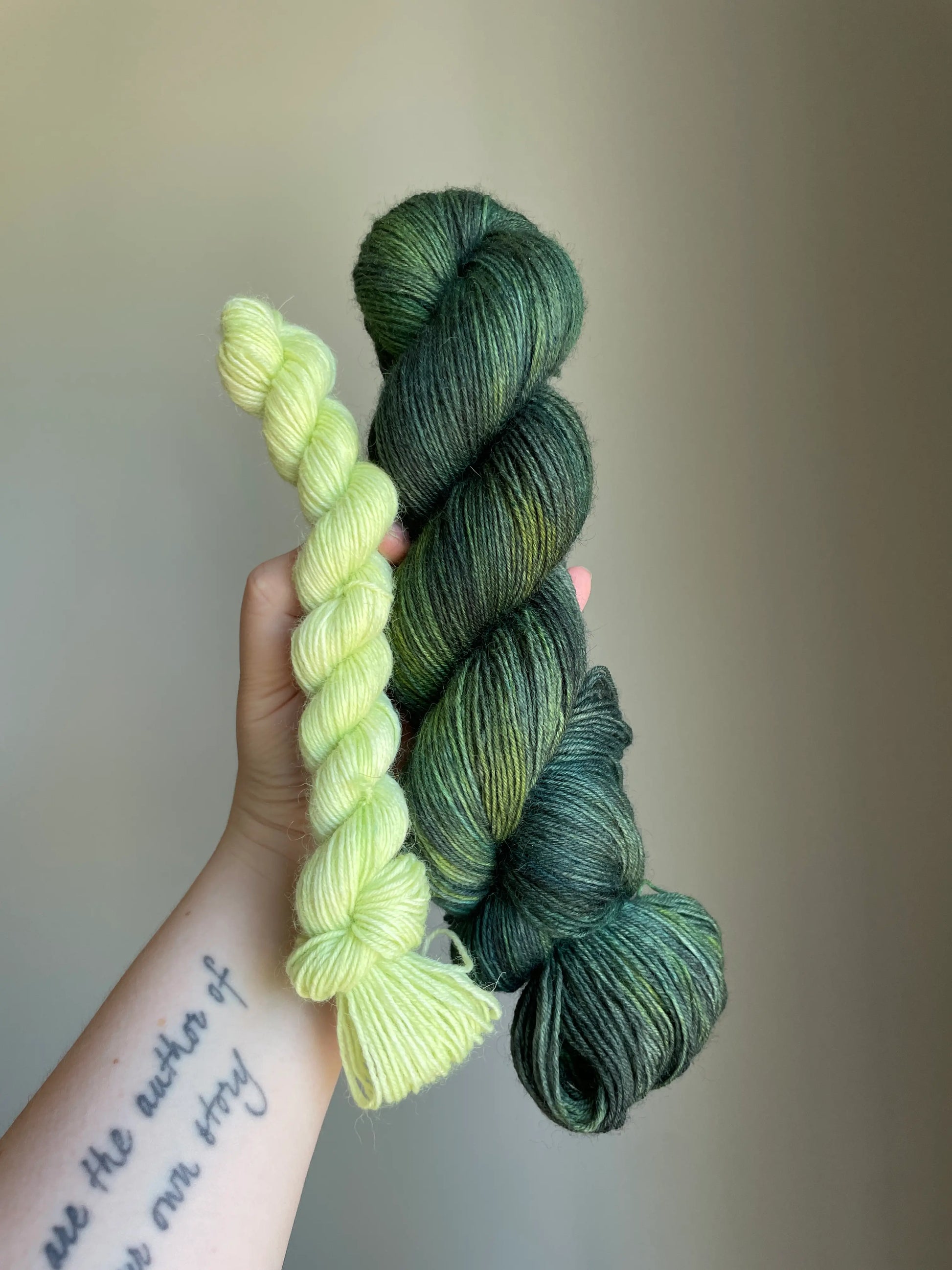 Handfärgat sockgarn-set med 100 g sockgarn i skogsgröna, petrolblå och mossgröna nyanser samt ett 20 g mini-nystan i ljus limegrön kontrastfärg. Garnset för stickade strumpor, sockor och accessoarer där mini-nystanet passar till häl, tå och mudd. Fotograferat på rustikt träbord som framhäver garnets färgskiftningar och glans.